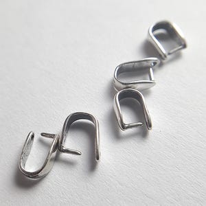 Puede incluir: Varias pequeñas argollas plateadas para colgantes se encuentran sobre una superficie blanca. Las argollas tienen un diseño en forma de U con un pequeño poste para sujetar un colgante o dije.