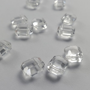 Op de afbeelding: Heldere, gefacetteerde kristallen kralen verspreid op een lichtgrijze ondergrond. De kralen hebben een rechthoekige vorm met gesneden randen, die licht reflecteren. Geschikt voor het maken van sieraden en knutselprojecten.