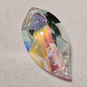 Peut inclure: Un pendentif en cristal clair en forme de feuille. Le cristal facetté réfracte la lumière, affichant un spectre de couleurs comprenant du jaune, du rose et du bleu. Le pendentif est posé sur un fond neutre, mettant en valeur ses coupes complexes et ses surfaces réfléchissantes.