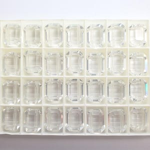 May include: A tray of clear, faceted, rectangular gemstones. Each gem is set in a white compartment. The gems have a reflective quality, catching the light and creating a sparkling effect. The tray is on a white surface.