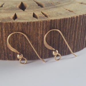 May include: A pair of gold-toned metal earring hooks featuring a curved wire design with a small spherical bead and a circular loop at the base for attaching jewellery components. The hooks have a smooth, polished finish and a simple, functional structure.