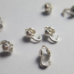 May include: A collection of small, silver-colored metal crimp covers, also known as bead tips or clamshells. Each piece features a rounded, hollow dome shape with an attached hook or loop for securing jewelry wire or cord ends in crafting projects.