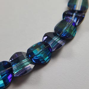 May include: Close-up of a strand of iridescent blue and green faceted beads. The beads are heart-shaped and have a reflective, multi-coloured surface. The beads are strung together, creating a curved line against a light grey background.