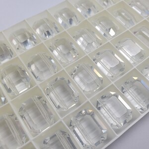May include: A tray filled with clear, faceted glass beads. Each bead is rectangular with cut edges, creating a sparkling effect. The beads are arranged in individual compartments within the white tray, suggesting a craft or jewelry-making supply.