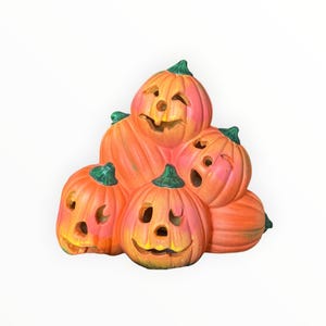 May include: A decorative ceramic sculpture featuring a stack of five orange pumpkins with green stems. Each pumpkin has carved facial features including eyes, noses, and mouths, creating a jack-o'-lantern appearance with a smooth, glossy finish.