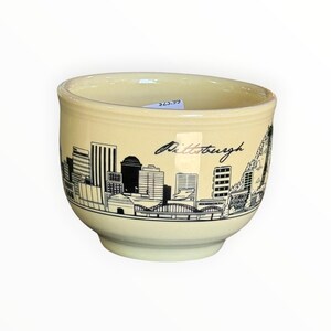 May include: A cream-colored ceramic bowl featuring a black line art illustration of the Pittsburgh city skyline. The design includes various buildings, a bridge, and the word Pittsburgh written in a cursive script across the front surface.