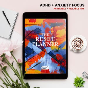 May include: A tablet displays "The Reset Planner" with an abstract art background in orange, blue, and white. The text "ADHD + Anxiety Focus" and "Printable + Fillable PDF" are at the top. A notebook, pen, coffee, flowers, and a succulent are also visible.