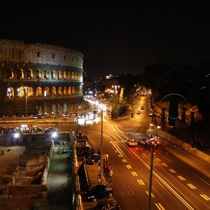 May include: A night view of the Colosseum in Rome, Italy, illuminated with lights. The ancient Roman amphitheater is surrounded by a street with cars driving on it.