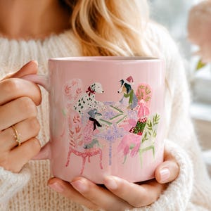 May include: A light pink ceramic mug featuring a colorful illustration of four dogs sitting around a table playing a game. The dogs are depicted wearing winter accessories like scarves and hats, set against a soft pink background on the mug surface.
