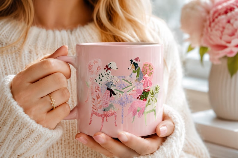 May include: A light pink ceramic mug featuring a colorful illustration of four dogs sitting around a table playing a game. The dogs are depicted wearing winter accessories like scarves and hats, set against a soft pink background on the mug surface.