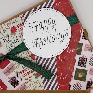 May include: A handmade Christmas card with a festive design. The card features a white circle with "Happy Holidays" in elegant script. The card is decorated with patterned paper in red, brown, and white, with a green ribbon. The card is a festive holiday item.