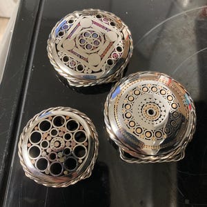 以下が含まれることがあります： 複雑なデザインのシルバーカラーのメタルベルトバックル3つ。1つはカラフルなアクセントのある幾何学模様、もう1つはゴールドカラーのディテールのある円形デザイン、3つ目は円形の切り抜きがあります。