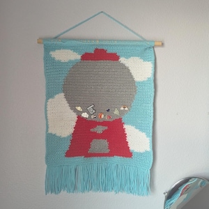 May include: A handmade crochet wall hanging featuring a gumball machine design. The artwork is primarily blue, gray, red, and white, with a fringe at the bottom. The gumball machine has small pins attached to it. The wall hanging is suspended from a wooden dowel.