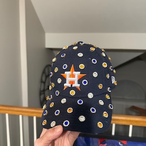 May include: A navy blue baseball cap with the Houston Astros logo, embellished with rhinestones. The cap features a white and orange embroidered logo and is adorned with clear, blue, and gold rhinestones.