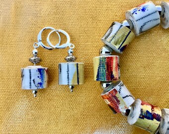 May include: A pair of silver hoop earrings and a beaded bracelet. The earrings and bracelet feature cylindrical beads with colorful abstract designs. The jewelry is set against a yellow background.