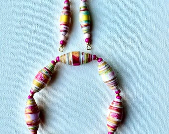 May include: A handmade jewelry set featuring a bracelet and earrings. The bracelet and earrings are made of colorful, cylindrical beads in shades of pink, yellow, and green. The earrings have gold-colored hooks. The beads are strung together with pink beads.
