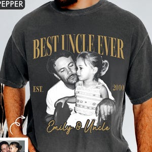 May include: Dark gray t-shirt with a sepia-toned photo of a man and child. The shirt reads "BEST UNCLE EVER" in gold lettering, with "EST. 2010" and "Emily & Uncle" below the photo. A small photo is in the corner.
