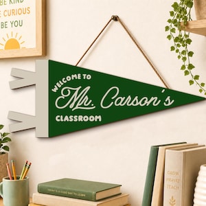 May include: A triangular green felt pennant hanging from a brown string. The pennant features white text that reads "WELCOME TO Mr. Carson's CLASSROOM" in a mix of block and script fonts. The left side has two white rectangular tabs.