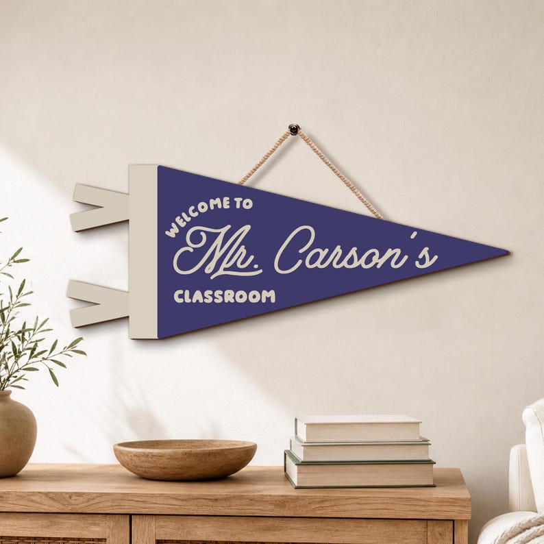 May include: A triangular dark blue wall pennant with a cream-colored header and two side tabs. The text "WELCOME TO Mr. Carson's CLASSROOM" is printed in cream-colored lettering. A tan rope is attached to the top for hanging the wall sign.