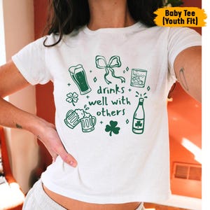 May include: White t-shirt with a green graphic design featuring beer glasses, a bow, a whiskey glass, a bottle, and shamrocks. The text "drinks well with others" is also included in the design. The shirt is labeled "Baby Tee (Youth Fit)".