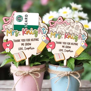 May include: Two wooden plant stakes with the text "Mrs. Knight" and "Thank you for helping me grow, Love, Sophia." Each stake has a pencil, apple, and book design. One stake holds a Starbucks gift card. The stakes are in pink and blue pots.