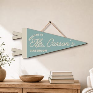 May include: A triangular wall pennant in light blue with a cream-colored side strip and two small tabs. The text reads "WELCOME TO Mr. Carson's CLASSROOM" in white lettering. A thin brown rope is attached to the top for hanging purposes.