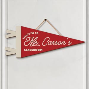 May include: A red triangular pennant banner with a cream-colored header and two side tabs. The text reads "WELCOME TO Mr. Carson's CLASSROOM" in cream-colored lettering. A tan rope is attached to the top for hanging the decorative wall sign.