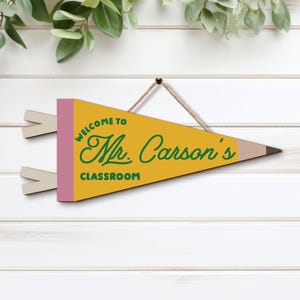 May include: A yellow pennant shaped like a pencil with a pink eraser end and a dark brown tip. The text reads "WELCOME TO Mr. Carson's CLASSROOM" in green lettering. The sign hangs from a thin brown cord attached to the top edge of the pennant.