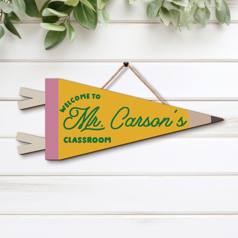 May include: A yellow pennant shaped like a pencil with a pink eraser end and a dark brown tip. The text reads "WELCOME TO Mr. Carson's CLASSROOM" in green lettering. The sign hangs from a thin brown cord attached to the top edge of the pennant.