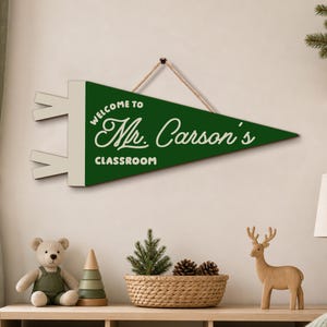 May include: A triangular green pennant wall sign with a cream-colored border and two tabs on the left side. The text reads "WELCOME TO Mr. Carson's CLASSROOM" in cream-colored lettering. A tan rope is attached to the top for hanging purposes.
