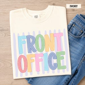 May include: An ivory t-shirt featuring the text FRONT OFFICE in multi-colored, scalloped-edge letters with vertical stripes. The letters are arranged in two rows over a background of light purple vertical stripes, paired with blue denim jeans.