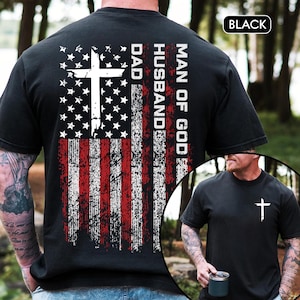 May include: Black t-shirt featuring a distressed American flag design with a white cross and the words "MAN OF GOD, HUSBAND, DAD." A smaller image shows a man wearing the same shirt with a white cross on the chest.
