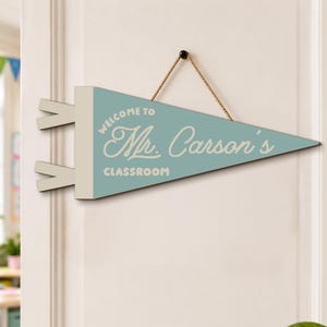 May include: A triangular light blue pennant sign with a cream-colored header and two side tabs. The text reads "WELCOME TO Mr. Carson's CLASSROOM" in cream-colored lettering. A brown twine string is attached to the top for hanging purposes.