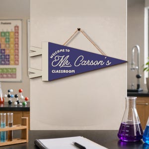 May include: A triangular dark blue pennant sign with a cream-colored border and hanging rope. The text reads "WELCOME TO Mr. Carson's CLASSROOM" in white lettering. The sign is oriented horizontally with a pointed end facing to the right side.