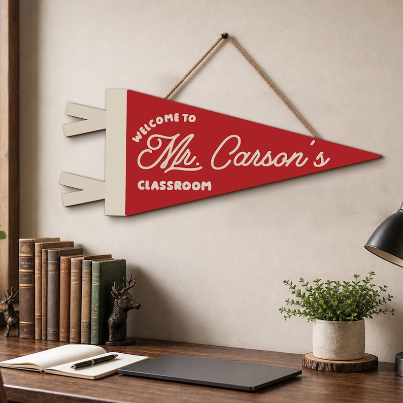 May include: A red and cream-colored triangular pennant sign featuring the text "WELCOME TO Mr. Carson's CLASSROOM" in white lettering. The pennant has a cream-colored header with two small tabs on the left side and hangs from a brown rope.