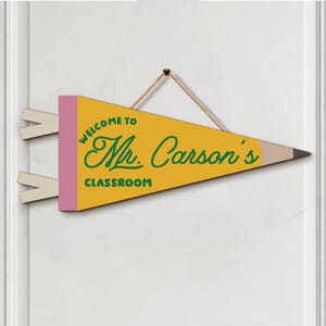 May include: A yellow, pencil-shaped pennant sign with a pink eraser end and a brown tip. The text reads "WELCOME TO Mr. Carson's CLASSROOM" in green lettering. The sign is suspended by a thin, light-colored rope attached to the top edge.