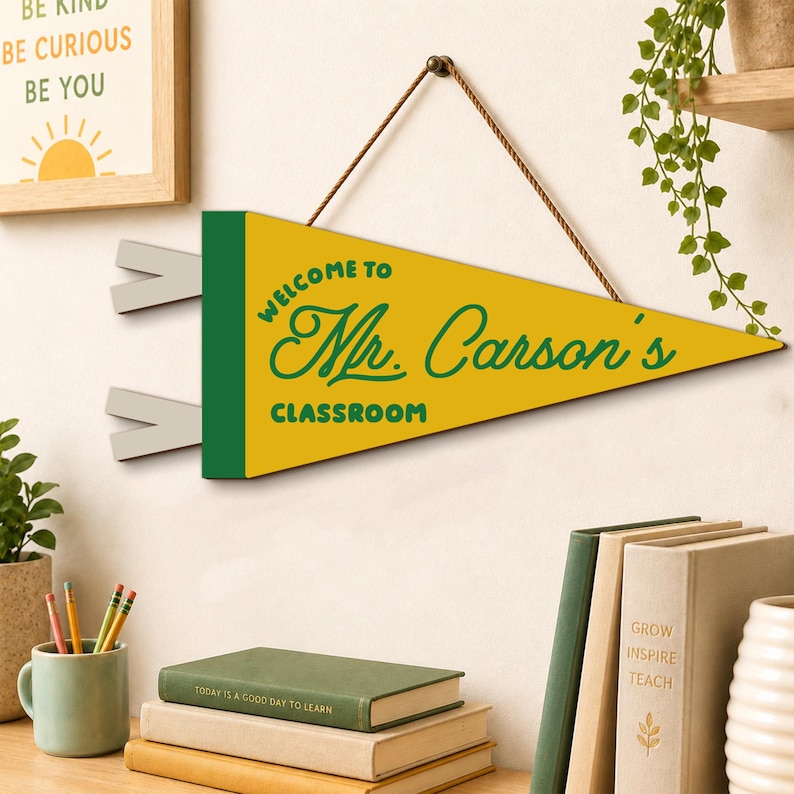 May include: A triangular yellow and green felt pennant hanging from a brown string. The text reads "WELCOME TO Mr. Carson's CLASSROOM" in dark green lettering. The pennant features two light gray fabric tails on the left side for attachment.
