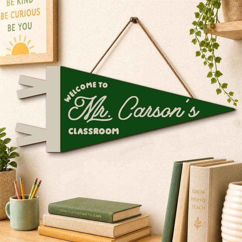 May include: A triangular green felt pennant hanging from a brown string. The pennant features white text that reads "WELCOME TO Mr. Carson's CLASSROOM" in a mix of block and script fonts. The left side has two white rectangular tabs.