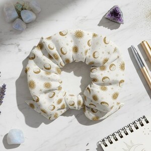 May include: White scrunchie with a gold celestial pattern of moons, stars, and suns. The scrunchie is surrounded by crystals, lavender, a gold pen, and a notebook with a sun and moon design.
