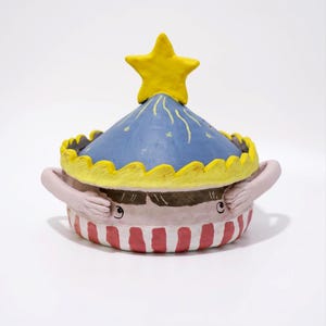 May include: A ceramic lidded bowl featuring a blue conical lid topped with a yellow star and yellow scalloped trim. The base is decorated with red and white vertical stripes and a face with brown hair and two hands reaching toward the lid.