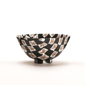 May include: A small bowl featuring a repeating pattern of black and white diamond shapes. Each white diamond contains a light brown, abstract, flower-like mark. The bowl has a rounded body and a small, circular base, with the pattern extending to the rim.