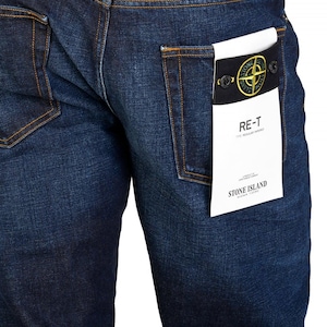 May include: Dark blue denim jeans with visible stitching and a back pocket. A white tag with black text and a logo is attached to the back pocket. The jeans are a regular tapered fit.