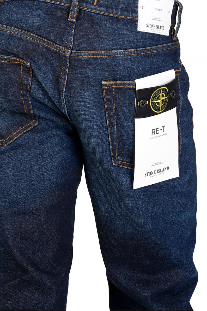 May include: Dark blue denim jeans with visible stitching and a back pocket. A white tag with black text and a logo is attached to the back pocket. The jeans are a regular tapered fit.