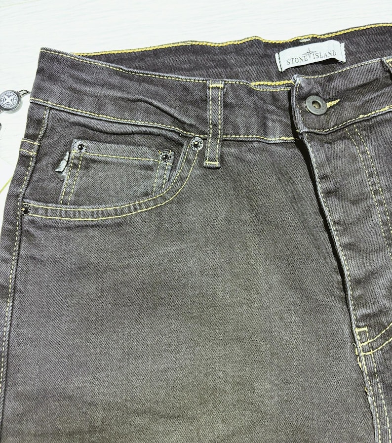 May include: Close-up of dark gray denim jeans with yellow stitching. The jeans have a classic five-pocket design, a button closure, and a small logo tag that reads "STONE ISLAND".