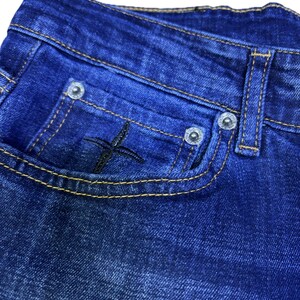 May include: Close-up of blue denim jeans. The jeans feature a small pocket with a black embroidered cross design. The pocket is outlined with yellow stitching. Silver rivets secure the pocket and belt loop.