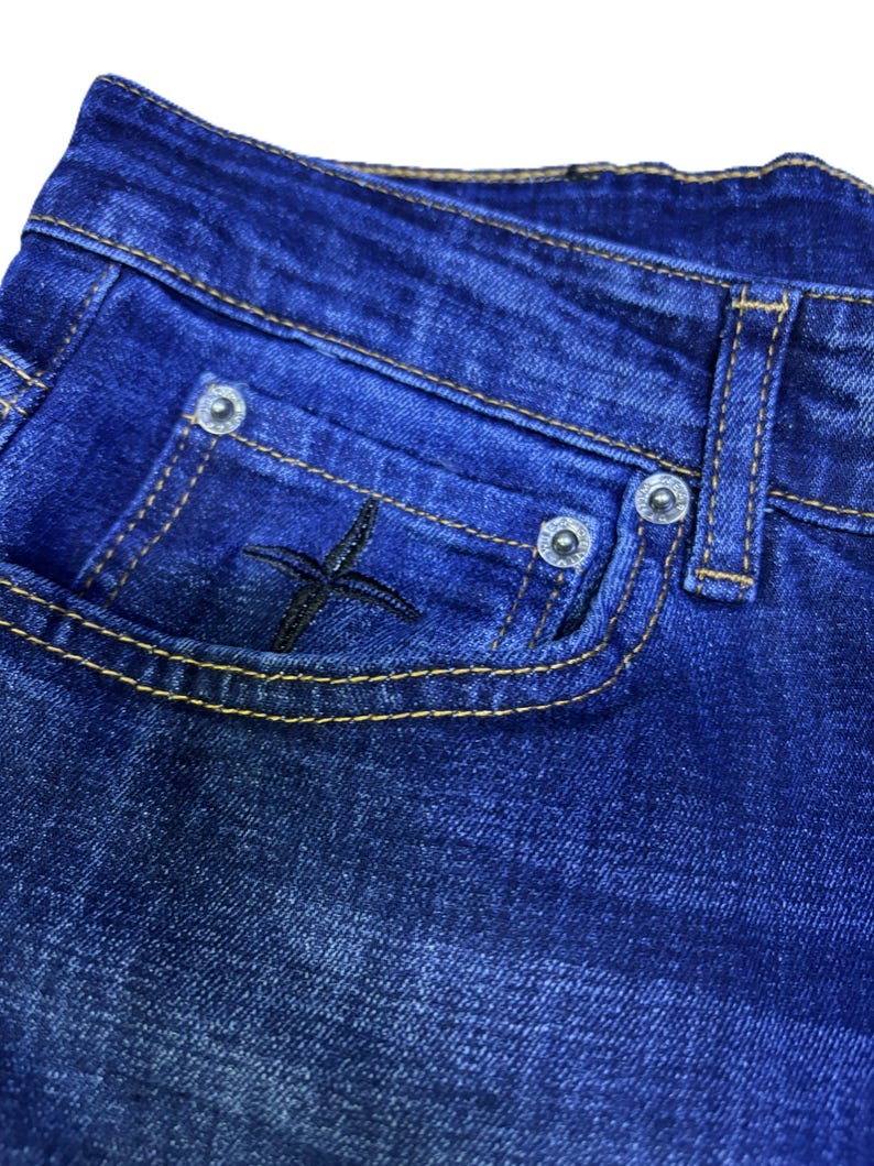 May include: Close-up of blue denim jeans. The jeans feature a small pocket with a black embroidered cross design. The pocket is outlined with yellow stitching. Silver rivets secure the pocket and belt loop.