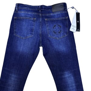 May include: Dark blue denim jeans with a faded wash. The back pockets feature a circular logo and a leather patch. A tag is attached to the jeans.