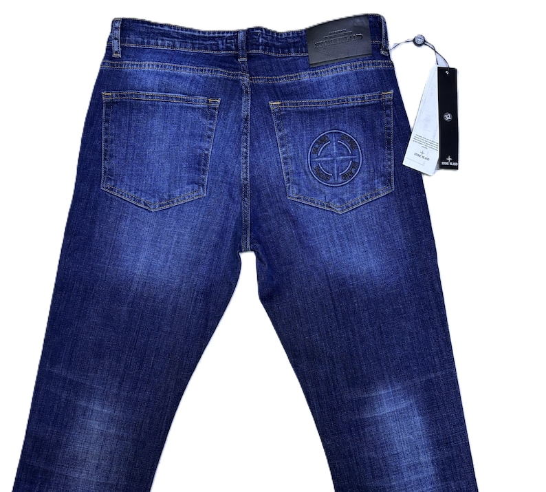 May include: Dark blue denim jeans with a faded wash. The back pockets feature a circular logo and a leather patch. A tag is attached to the jeans.