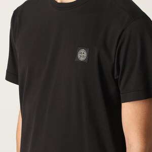 Puede incluir: Camiseta negra de cuello redondo y manga corta. Un pequeño parche cuadrado con un logotipo circular está en el pecho izquierdo. La camiseta está hecha de un material suave y de color sólido.