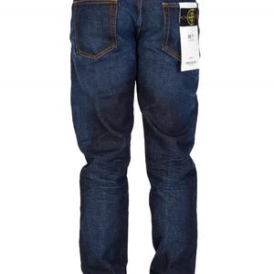 May include: Dark blue denim jeans with a classic five-pocket design. The jeans have a straight leg fit and a faded appearance. A white tag with black text is attached to the back pocket. The jeans are worn with white sneakers.